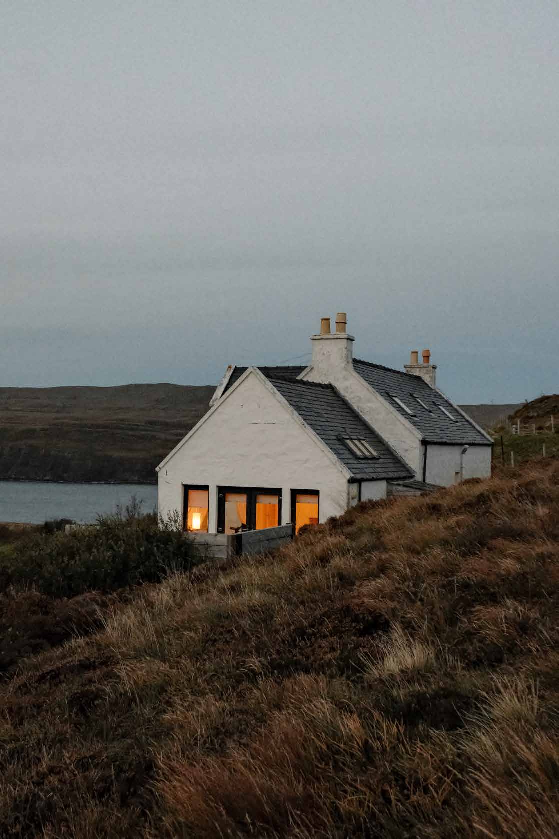 A white crofter's cottage on a hillside above a sea loch at dusk, its windows lit